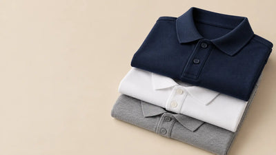 Three folded blank pique polo shirts in navy, white and heather grey stacked to show their collars and button plackets, on a warm beige studio background