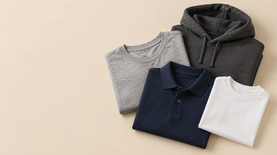 Folded blank grey t-shirt, navy polo shirt, charcoal hoodie and small white youth t-shirt arranged together on a warm beige studio background