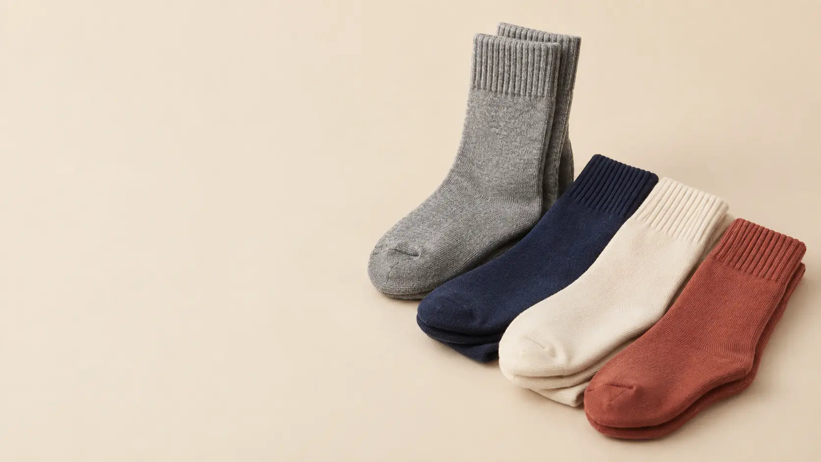 Four pairs of folded plain crew socks in heather grey, navy, cream, and terracotta arranged on a warm beige studio background